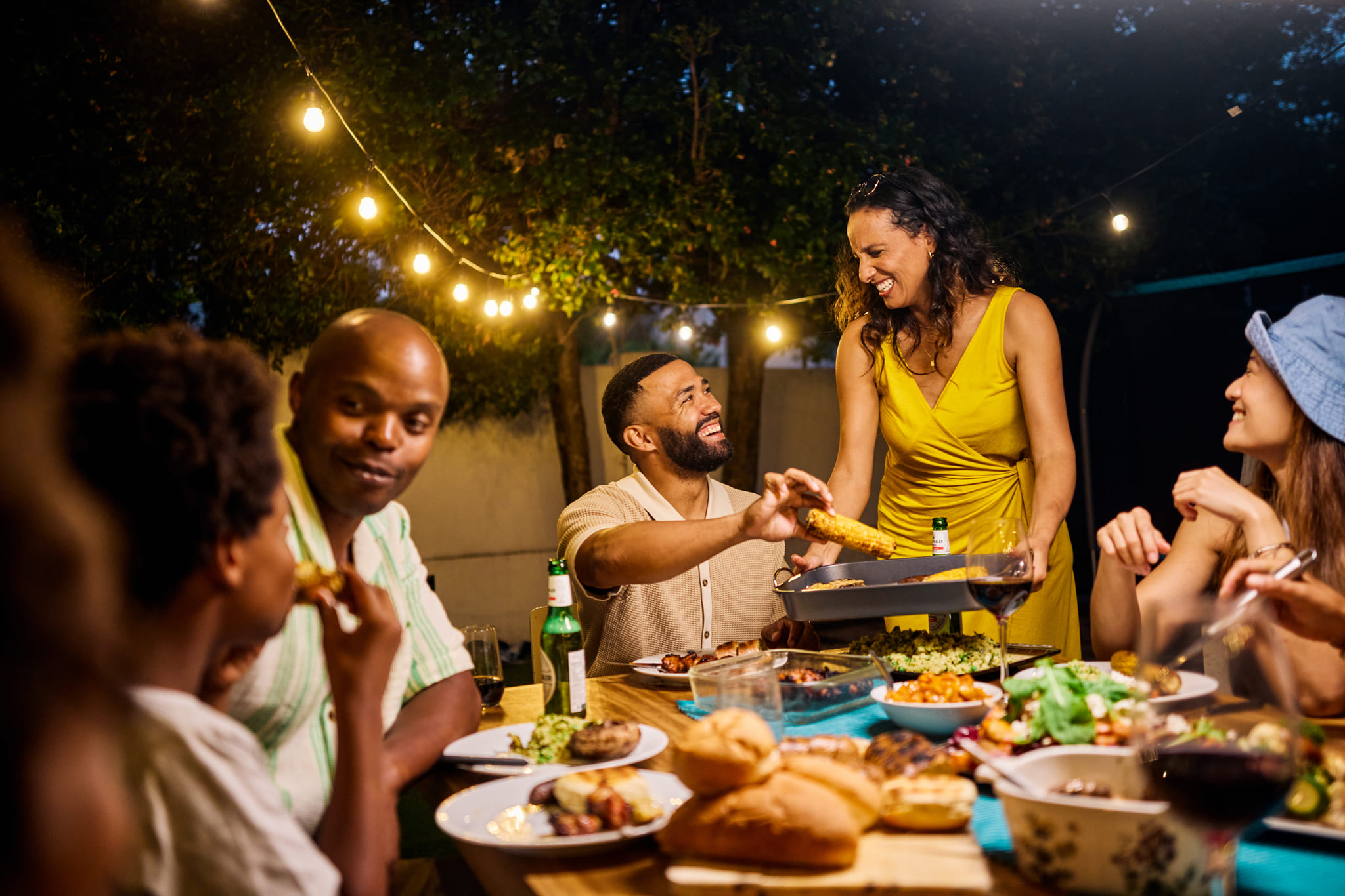A group of friends laughing and sharing food at an outdoor dinner party at night